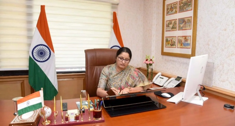 Annpurna Devi signs a file at a desk flanked by two national flags, with framed paintings on the wall
