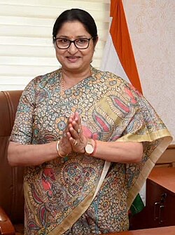 Official portrait of Annpurna Devi, hands folded in greeting, in a Madhubani-print sari beside the national flag