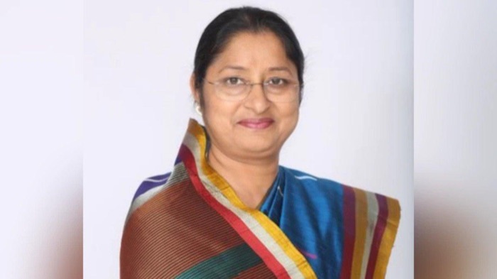 Portrait of Annpurna Devi, smiling, in spectacles and a striped multicoloured silk sari, against a plain pale background