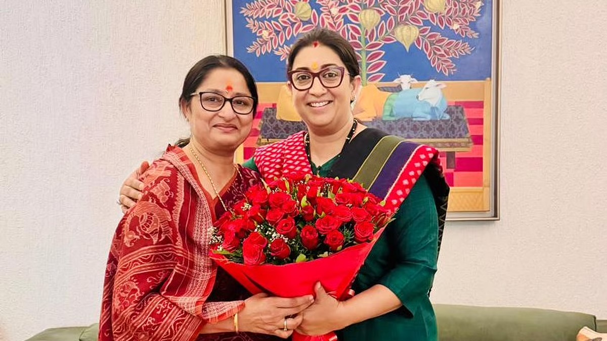Annpurna Devi and Smriti Irani stand together holding a bouquet of red roses in front of a painting