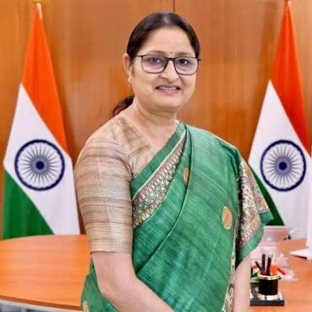 Annpurna Devi, smiling, in a green silk sari, standing in an office between two national flags