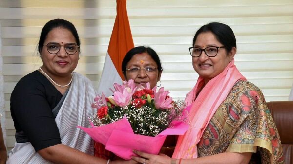 Annpurna Devi, smiling, receives a bouquet of pink lilies from two women colleagues in an office with the national flag