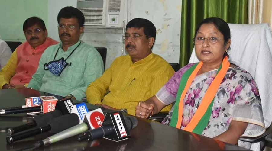 Annpurna Devi, wearing a party stole, speaks to the media from behind a table of news-channel microphones, seated beside three colleagues